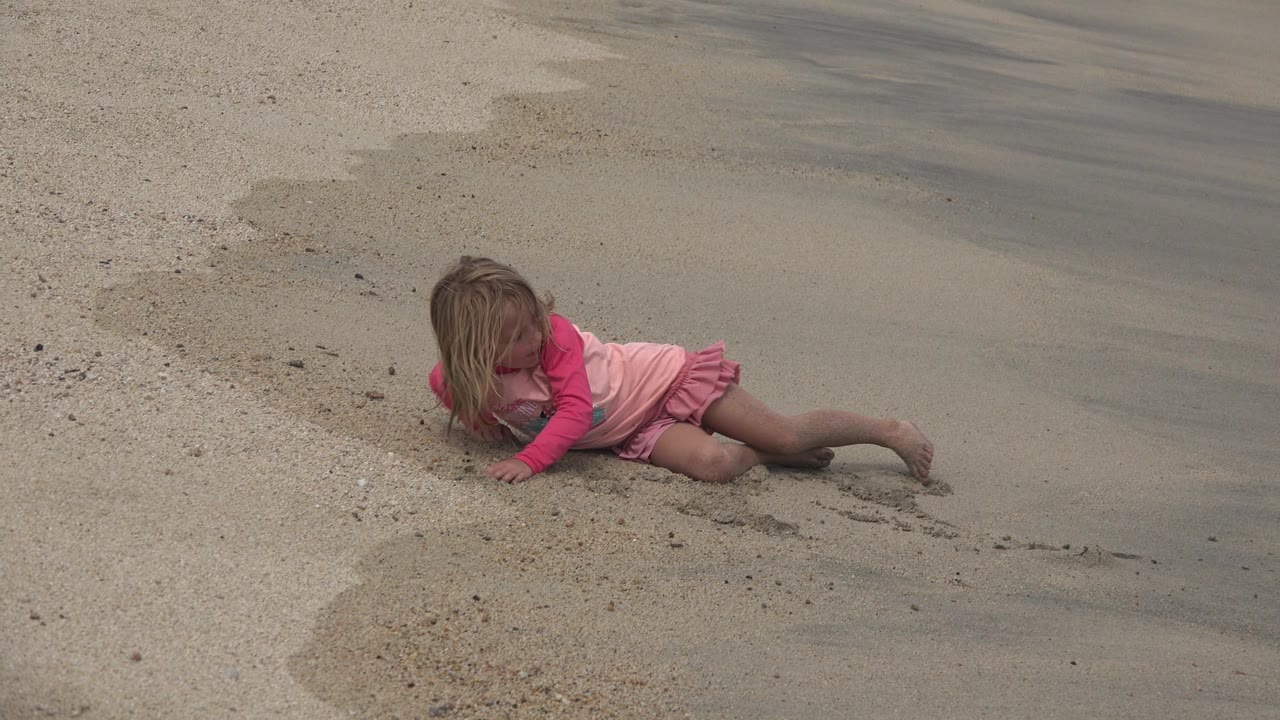 At 0:01, the child remains on the sloped sand in San Jose del Cabo, Mexico, facing partly downward with one arm pressed into the beach and wet sand behind her.