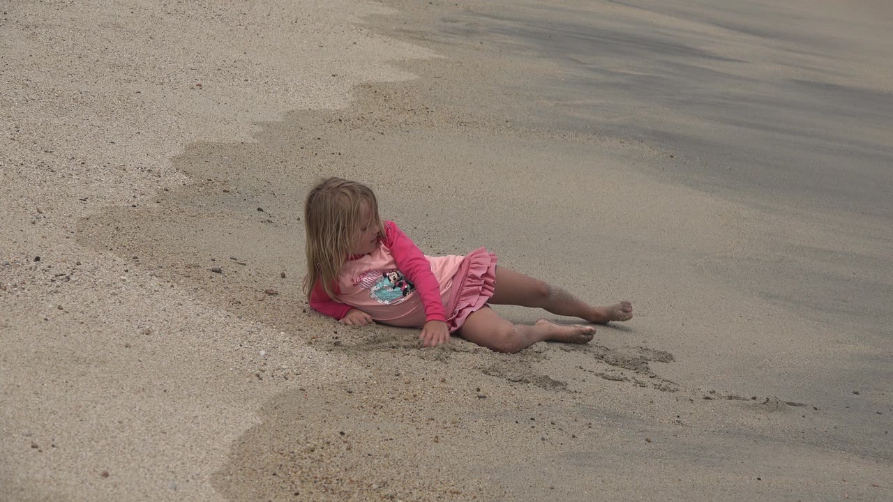 At 0:02, the girl sits more upright on the San Jose del Cabo beach, legs extended across the sand as she looks toward the wet shoreline.