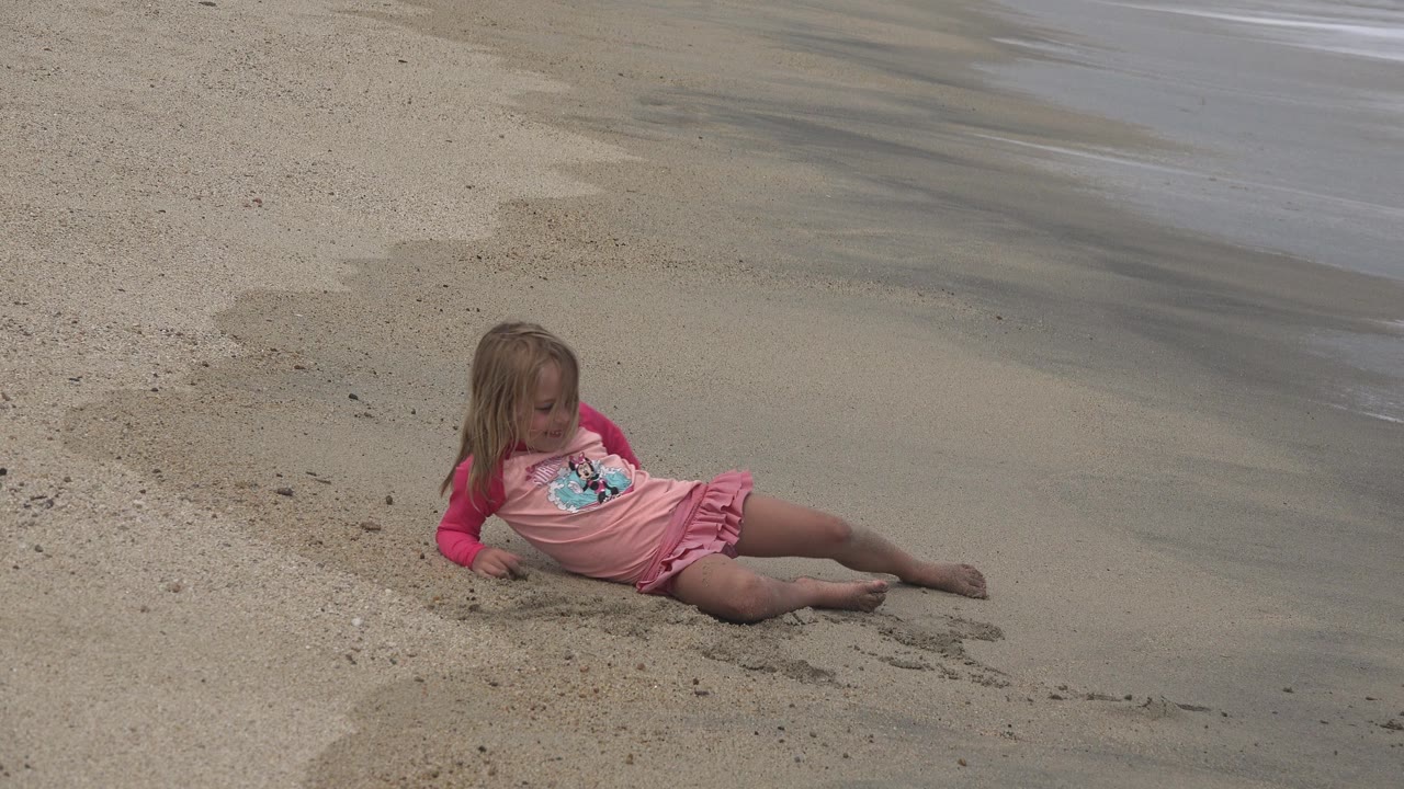 Young girl in a pink shirt sitting on tan beach sand near shallow water.