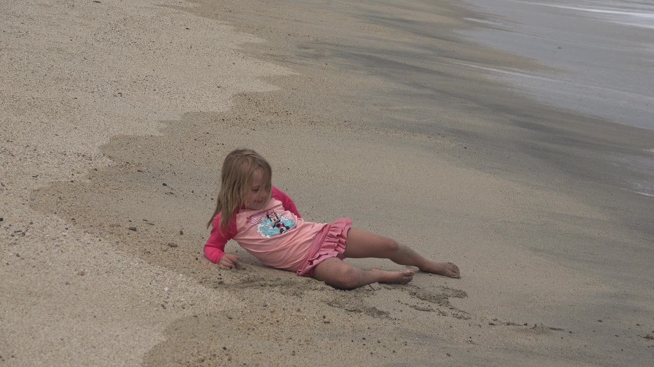 Young girl in pink remains seated on tan beach sand in San Jose del Cabo, Mexico, with one leg stretched toward the wet sand.