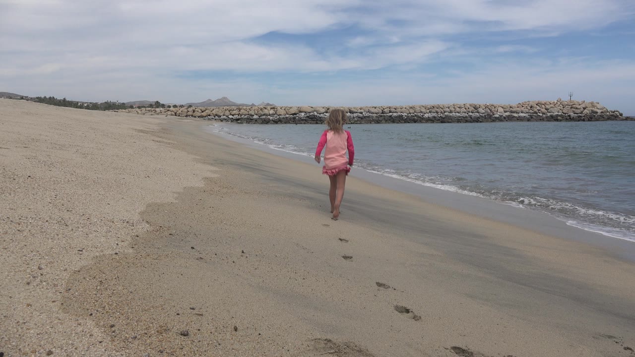 The girl continues along the San Jose del Cabo shoreline, centered against pale sand, blue-gray water, and the long rock barrier across the horizon.