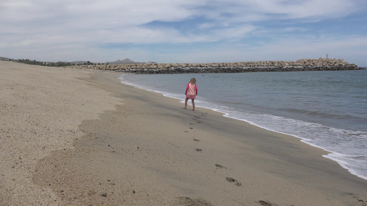 Young girl in pink standing near small waves on a sandy beach.