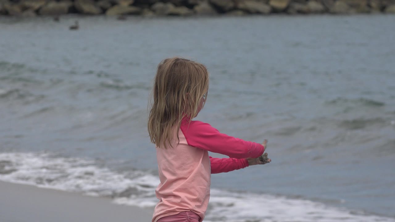 A young girl in a pink beach outfit faces the water in San Jose del Cabo, Mexico, with small waves and a rock breakwater behind her.
