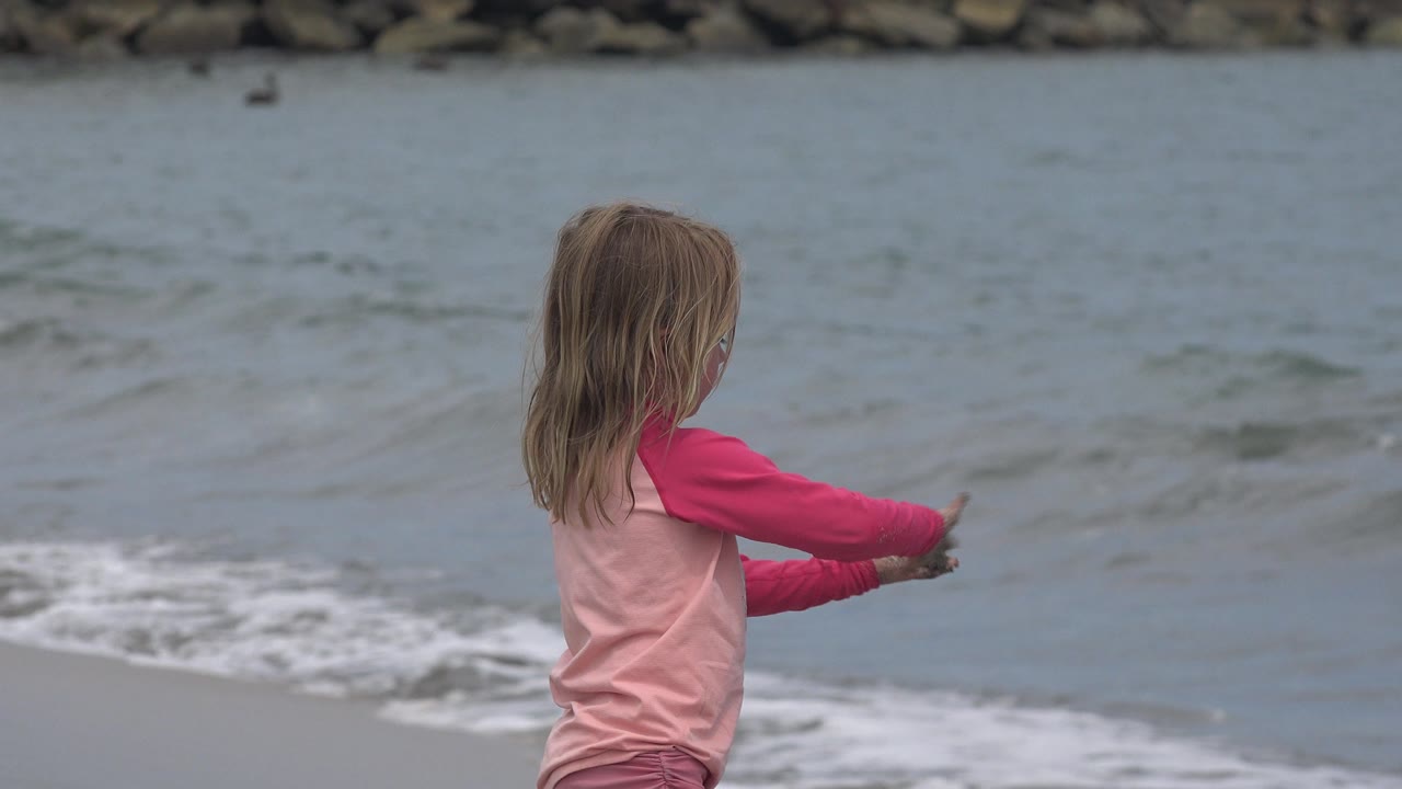 A young girl in pink stands in profile at the San Jose del Cabo shoreline, holding sand or small debris while a wave rolls in behind her.