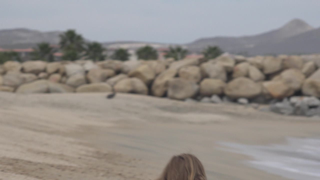 A second low frame in San Jose del Cabo shows the child's head barely entering the bottom of the shot, with pale sand, a rock barrier, scattered trees, and hazy hills behind.