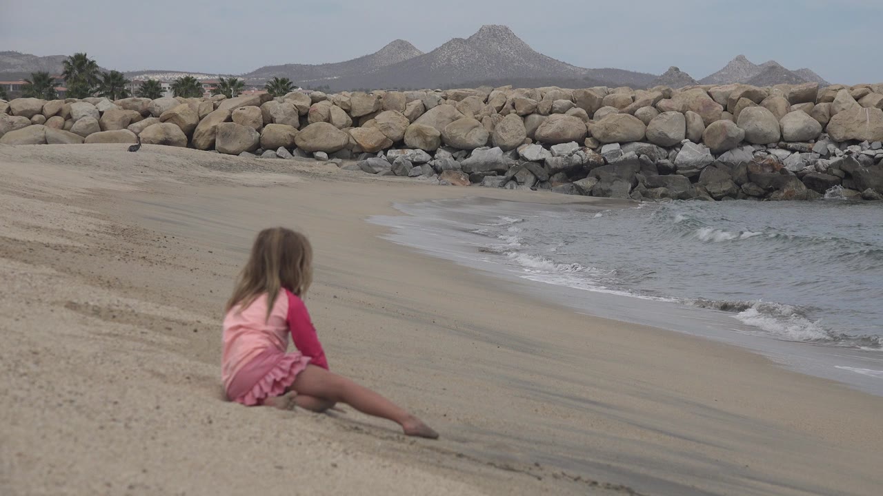 At 0:00, a young girl in a pink outfit sits low on the sand near the waterline in San Jose del Cabo, Mexico, with a rock breakwater and dry hills behind her.