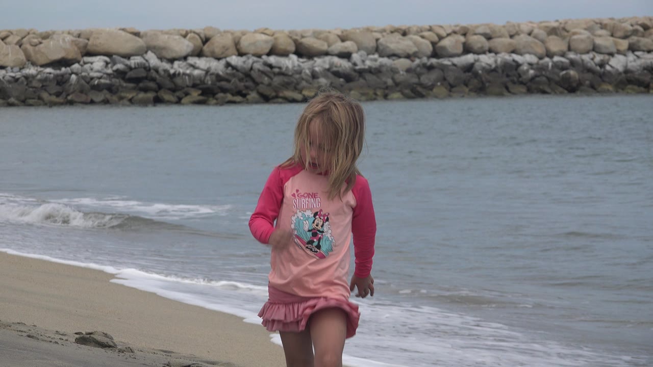 Young blonde girl in a pink dress walking on wet sand by the water.