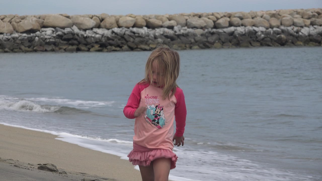 A young blonde girl in pink remains near the foreground as a low wave curls along the San Jose del Cabo shoreline and the stone seawall stretches across the background.