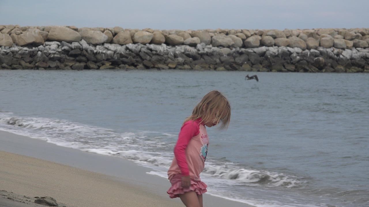 A young child in a pink long-sleeve beach outfit walks near small waves on a sandy beach in San Jose del Cabo, Mexico, with a stone breakwater across the water behind her.