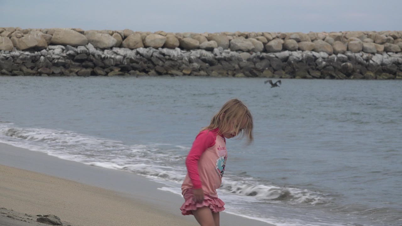 The child looks down toward the sand while walking along the surf in San Jose del Cabo, Mexico, with gray-blue water and the rock breakwater filling the background.