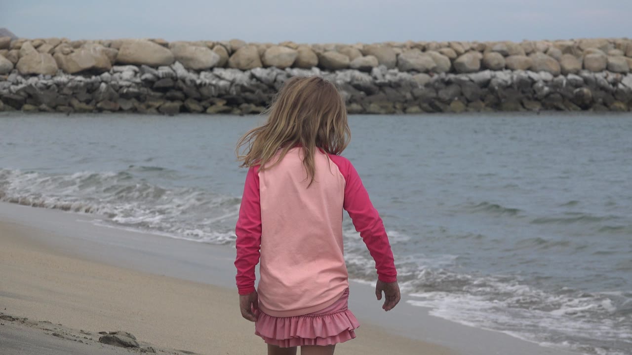 Child in pink walking near the surf with a rock breakwater behind.