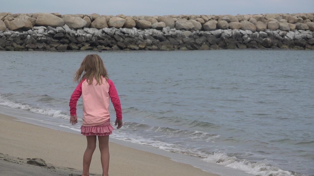 At 0:00, a young girl in pink stands with her back to camera near the surf on a San Jose del Cabo beach, with a rock breakwater across the water.