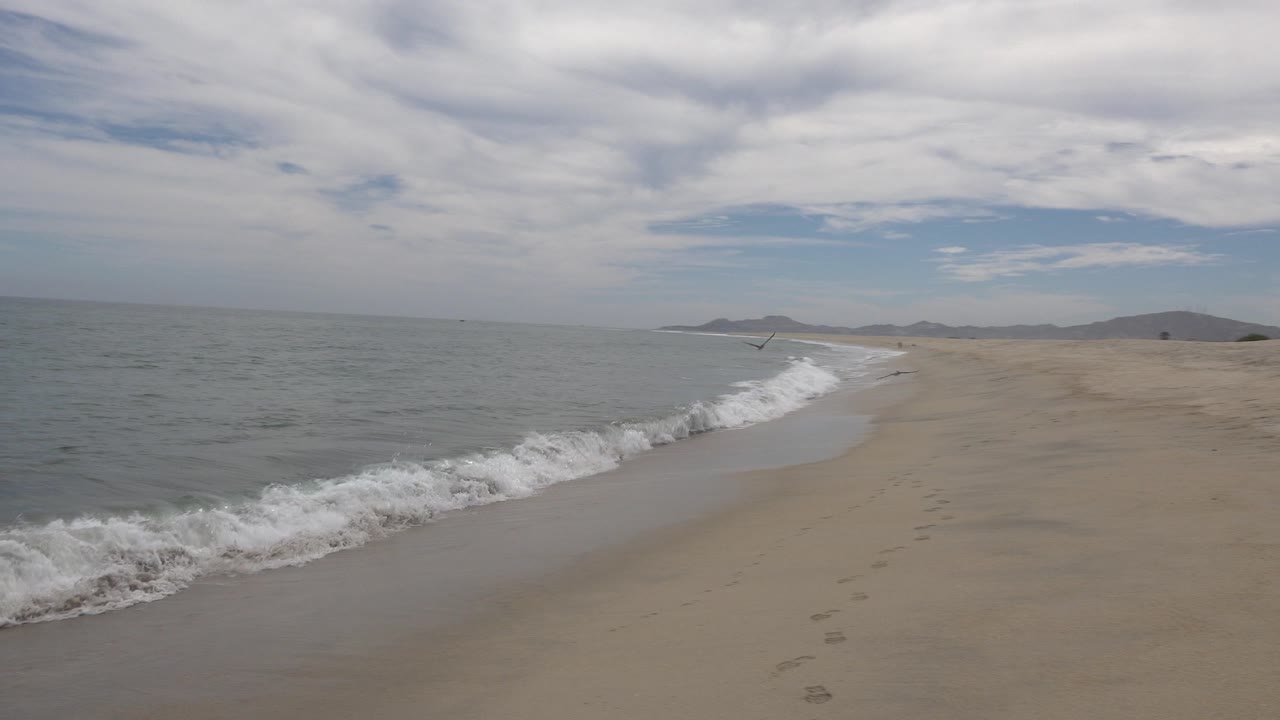 San Jose del Cabo beach at 0:01 with a fresh band of white foam curling along the diagonal shoreline, open water to the left, broad sand to the right, distant hills, and no visible people.