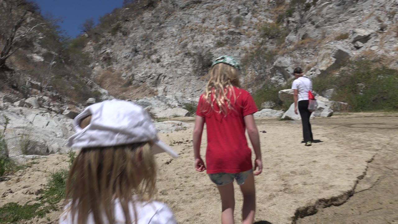 In San Jose del Cabo, two children are seen from behind in the foreground while an adult walks ahead on a sandy path below pale rocky slopes.