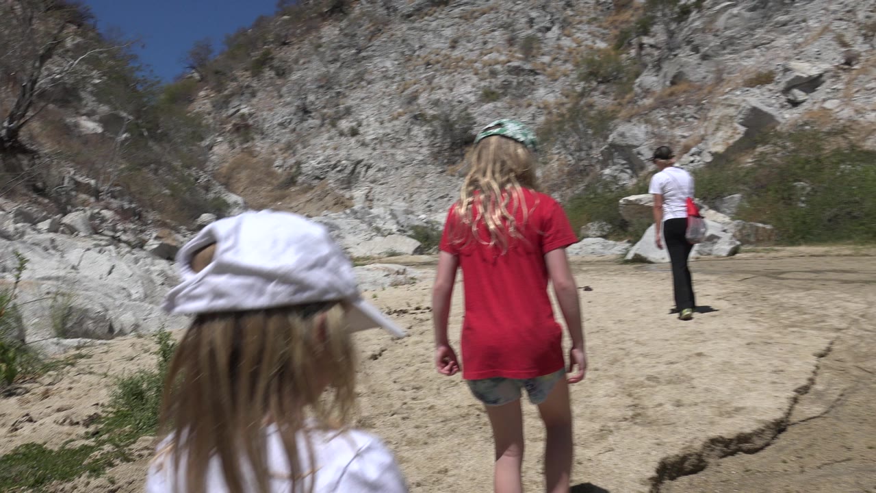 A child in a white cap fills the foreground while another child in a red shirt and an adult continue along the sandy San Jose del Cabo path.