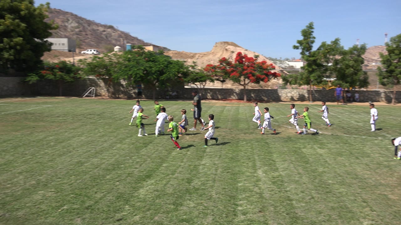 Players in white and neon green chase the soccer ball across the grass in San Jose del Cabo, Mexico, with the goal set back at the left and hills in the distance.
