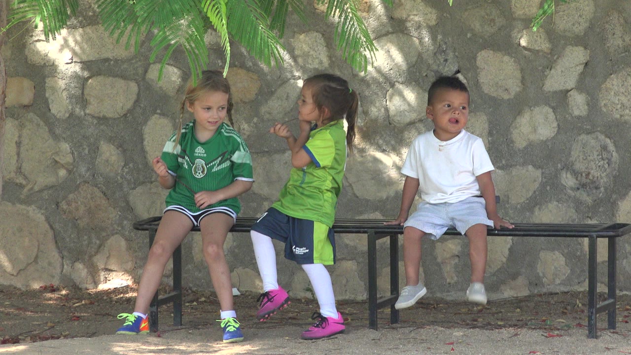 Three children are beside a black bench in San Jose Del Cabo, Mexico, with two girls in green soccer shirts at left and a boy in white seated at right.