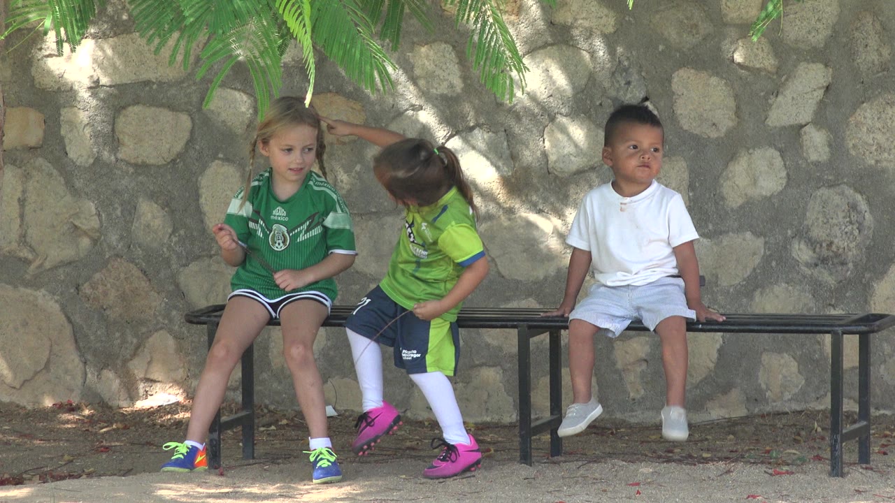 Girl in green touches another girl's head near a bench.