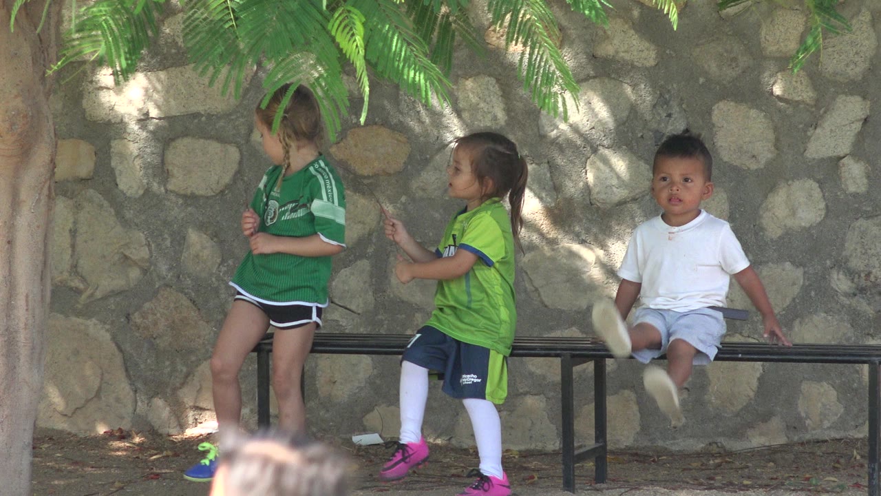 At one second, the same three children wait near the bench in San Jose del Cabo, Mexico, while the seated child in white moves his legs and the two children in green stand nearby.
