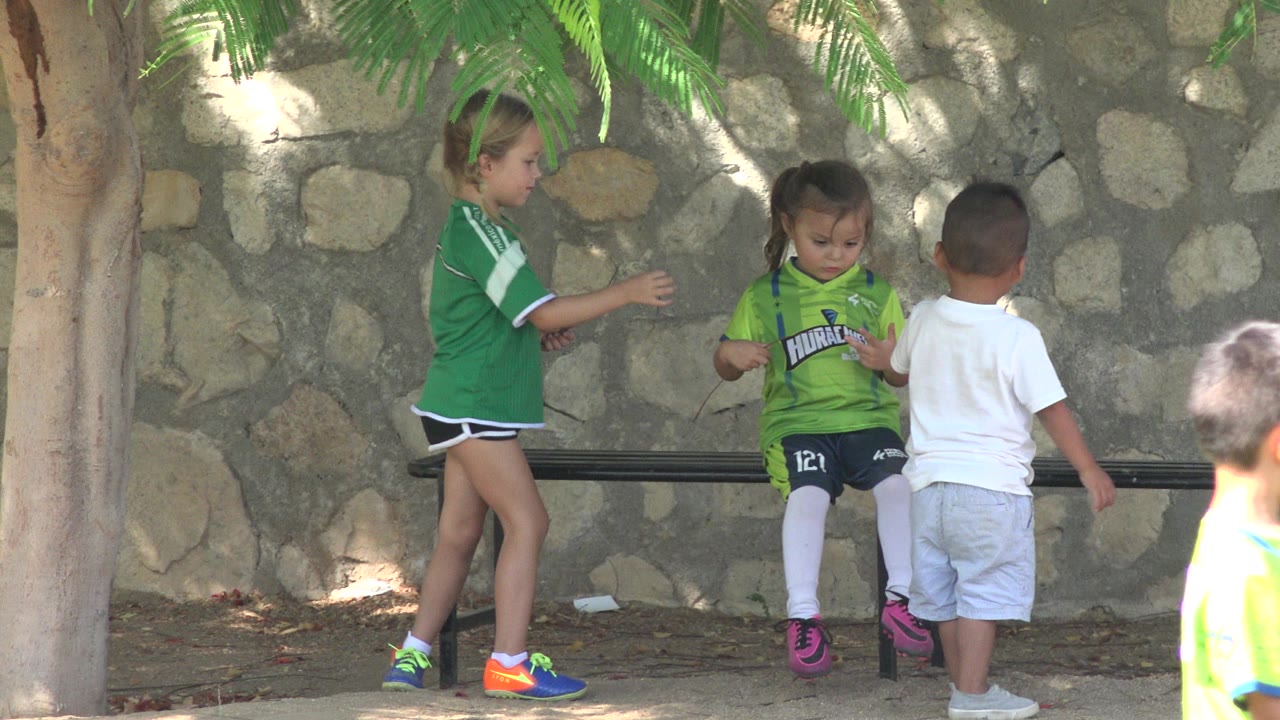 In San Jose Del Cabo, Mexico, the seated girl looks down at her hands while another girl stands to the left and a boy faces the bench in front of her.