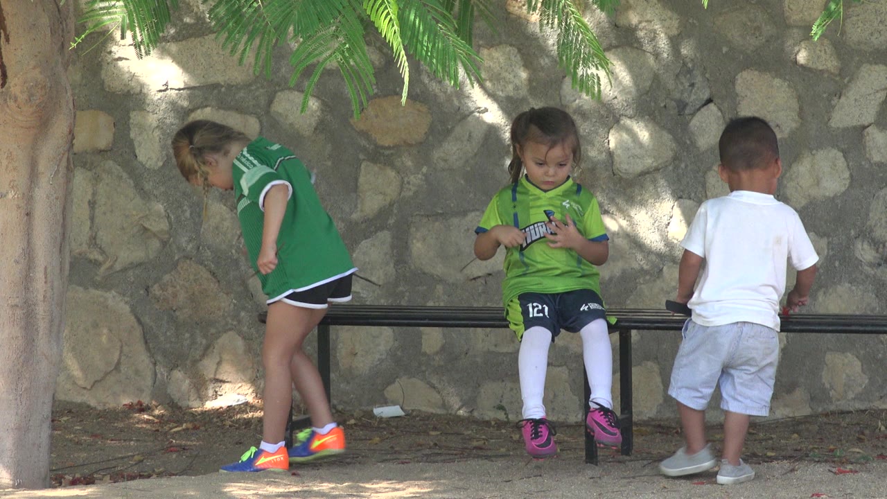 At 0:01 in San Jose del Cabo, Mexico, a child sits on the bench looking down while another bends near the left side and a boy in white faces away.