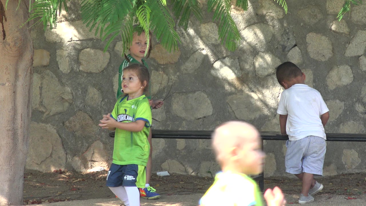 Two children in green soccer shirts face different directions while a third child in a white shirt stands by the bench against the San Jose del Cabo stone wall.