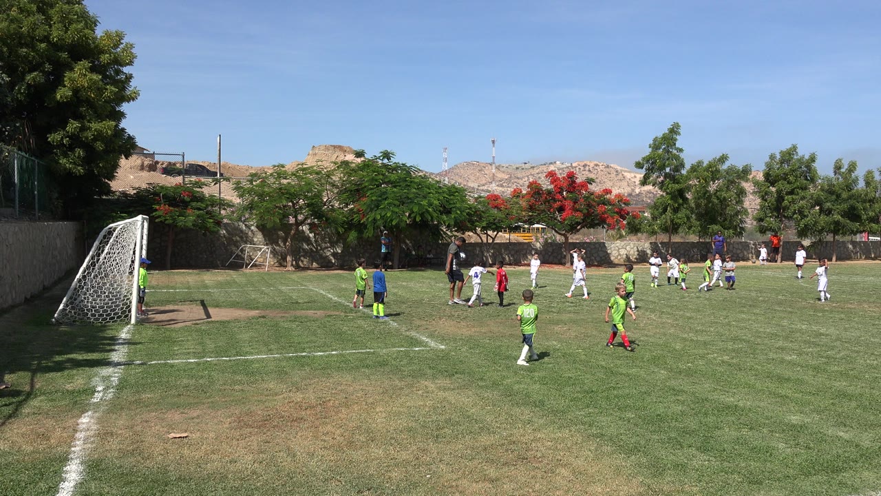 Wide frame of a youth soccer field in San Jose Del Cabo, Mexico, with children in green and white uniforms near a goal and a coach standing by the ball.