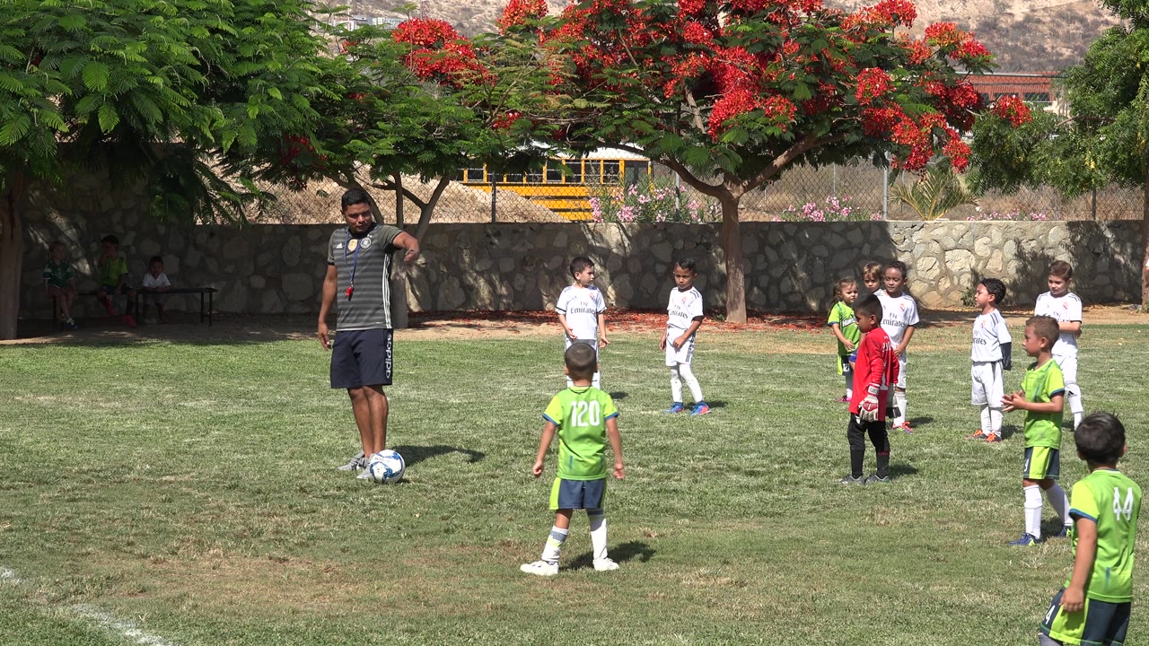 A coach stands with one hand near his hip while children cluster across the midground of a sunny soccer field in San Jose Del Cabo, Mexico.