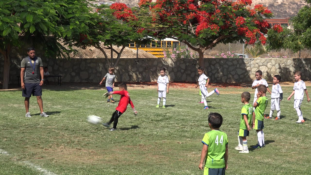 A boy in a red top swings his leg toward the soccer ball on the San Jose del Cabo school field as children in green and white uniforms stand nearby under bright sun.
