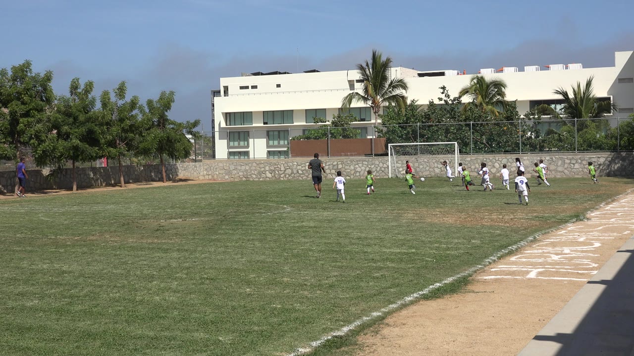 Children in white and green football uniforms play near a goal on a grass field in San Jose del Cabo, Mexico, with trees and white buildings behind the fence.