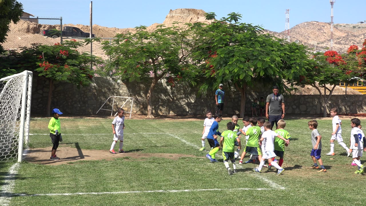 Kids in green and white uniforms chase the ball in front of the goal in San Jose Del Cabo, Mexico, while an adult stands near the penalty area.