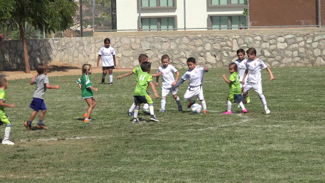 A group of children in white and green uniforms press toward the soccer ball on a sunny field in San Jose del Cabo, Mexico.