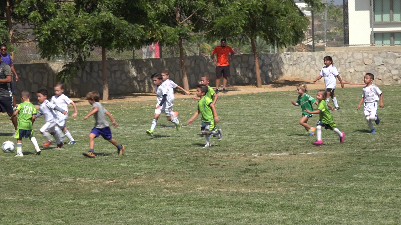 Children in mixed white and green uniforms sprint across the grass in San Jose del Cabo, Mexico, with a coach-like adult and stone wall behind them.