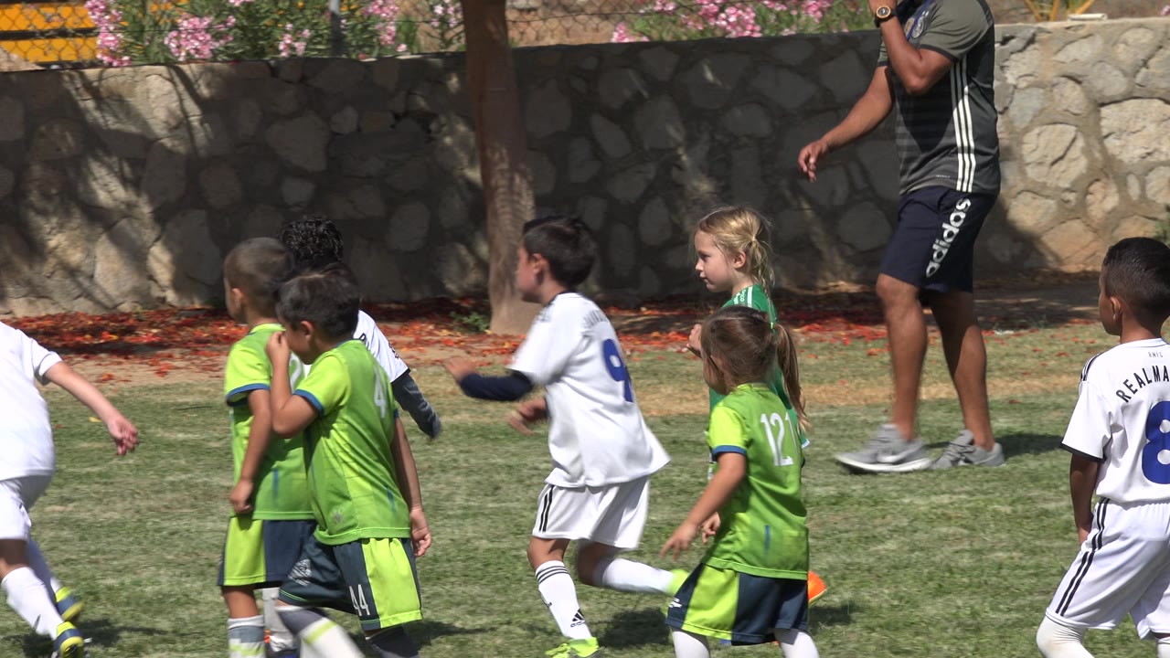 Children in green and white kits pass in front of an adult supervisor on a grass soccer field in San Jose del Cabo, Mexico, with partial faces and jersey numbers visible.