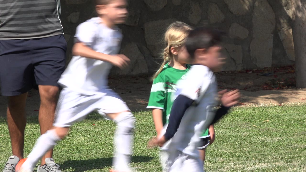 Children in soccer uniforms run past a girl in green on a grass field.