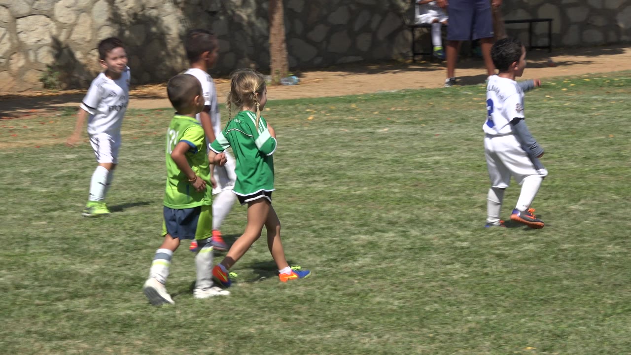 Children in green and white soccer uniforms walk and run on a grass field.