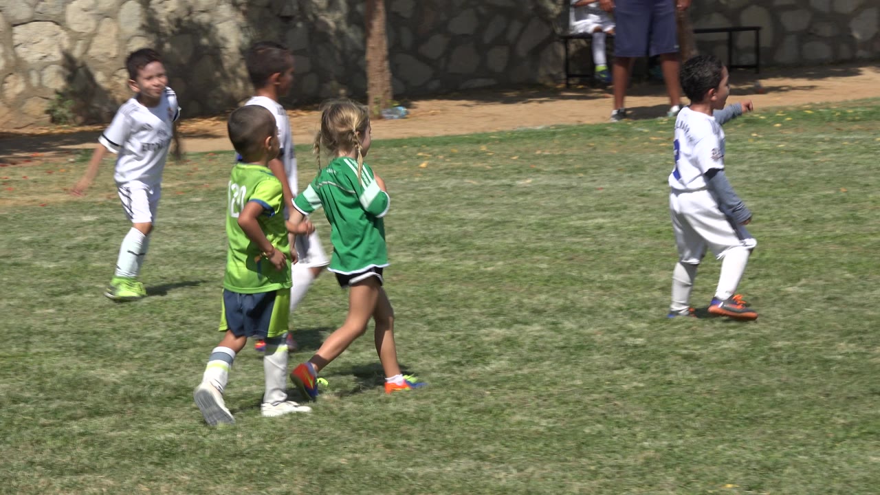 Young players in green and white uniforms continue across the grass in San Jose Del Cabo, Mexico, with the sideline wall and trees in the background.