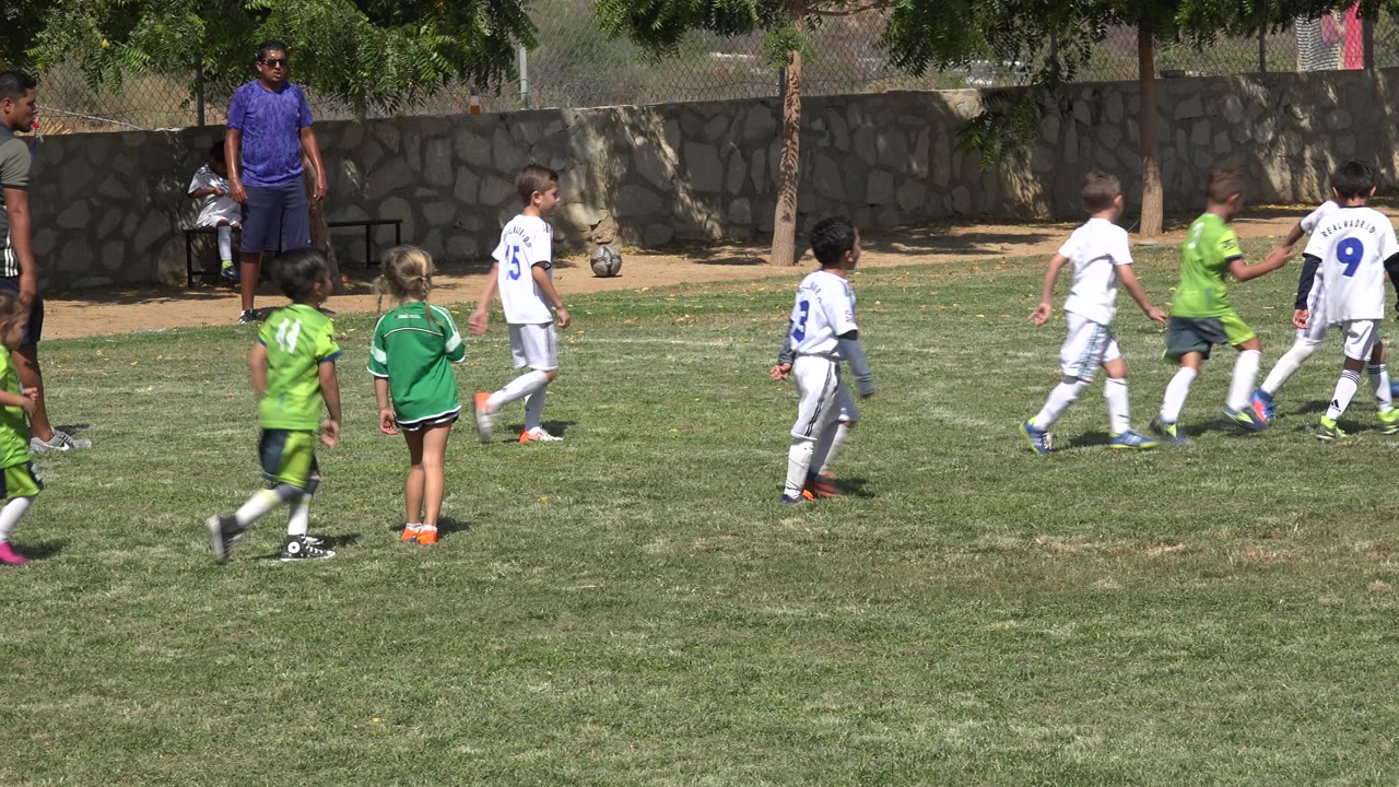 A group of children in green and white soccer kits move across the San Jose Del Cabo field while adults stand near the shaded sideline and stone wall.