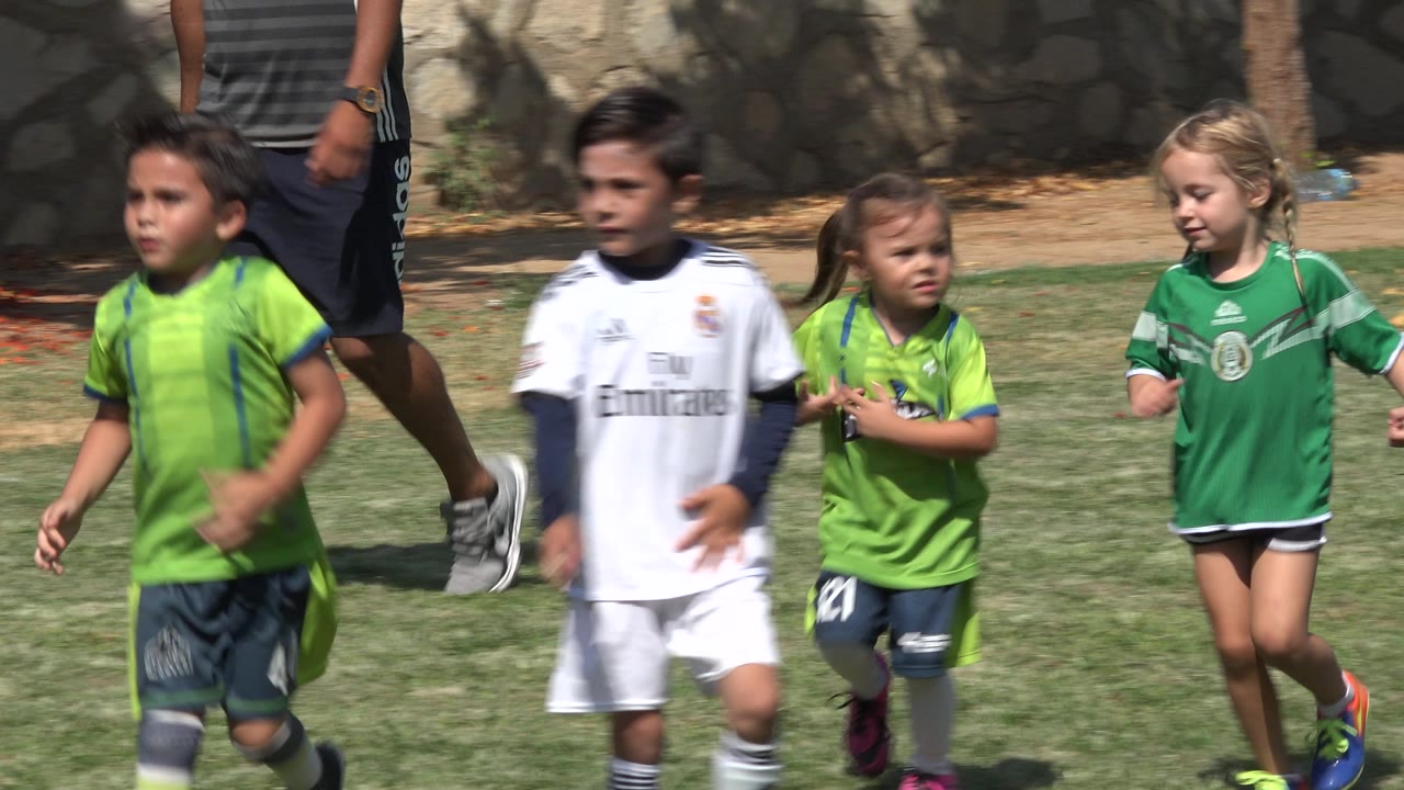 Children in green and white soccer jerseys jog across a grass field in San Jose Del Cabo, Mexico, with an adult's legs visible behind them near a stone wall.