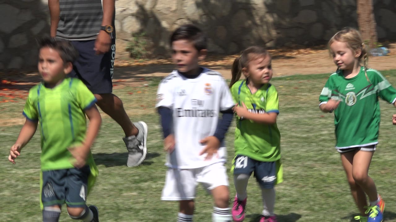 Several children in green and white soccer jerseys move through the frame on a sunny grass field in San Jose Del Cabo, Mexico, with the adult supervisor still partly cropped behind them.