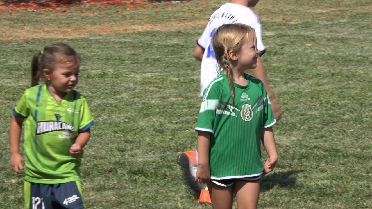 A young girl in a bright green soccer jersey stands in the foreground on a dry grass field in San Jose del Cabo, Mexico, while a child in white is partly visible behind her.