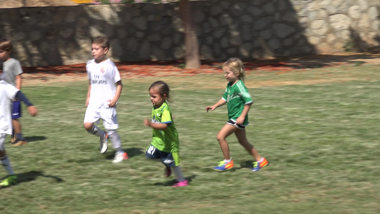Several children jog across the sunlit grass in San Jose Del Cabo, Mexico, including a child in a green uniform and children in white uniforms.