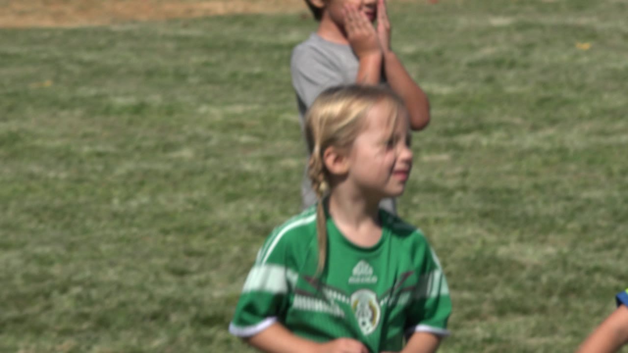 Blonde girl in green soccer jersey on grass field with child behind her.