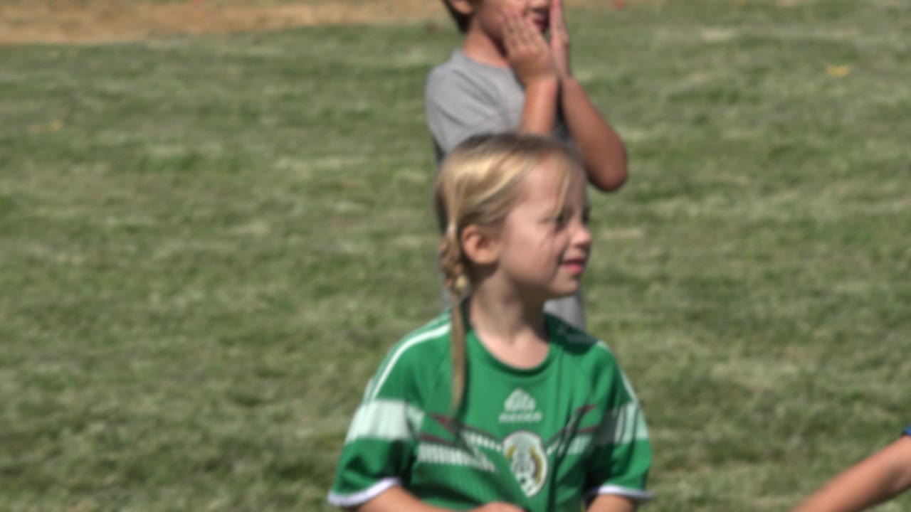 The girl in a green soccer jersey looks to the side on the sunlit field in San Jose Del Cabo, Mexico, while another child stands out of focus behind her.