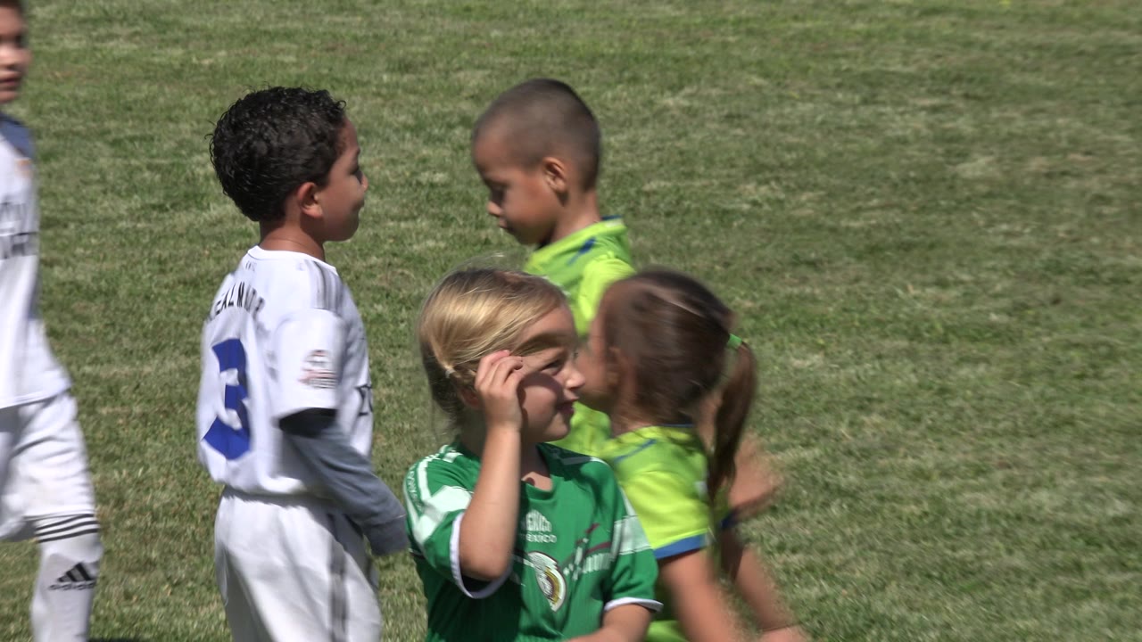Two children in white kits face a child in a bright green jersey while a blond child stands in front on the grass field in San Jose del Cabo, Mexico.