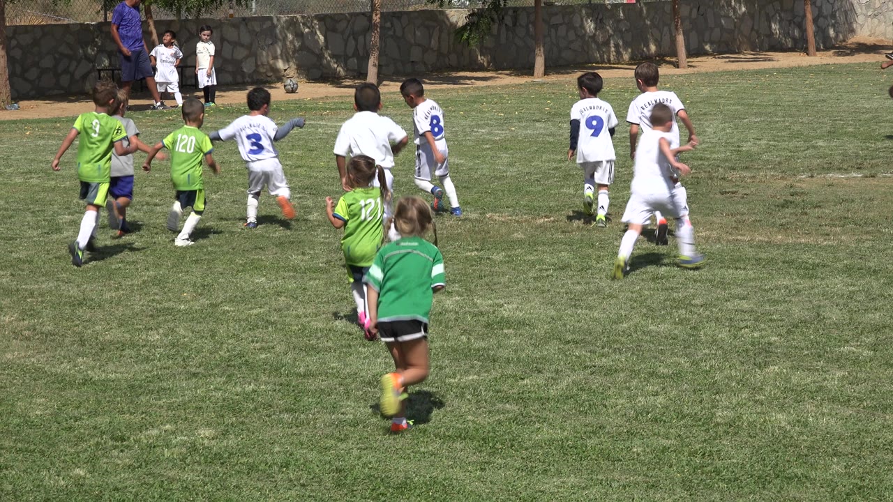 Children in green and white soccer uniforms run toward play on a grass field in San Jose del Cabo, Mexico, with a stone wall and trees behind them.