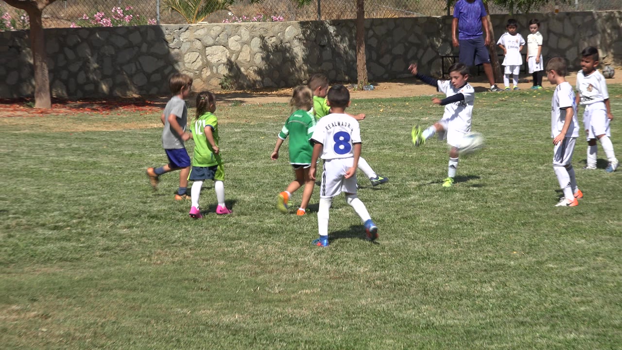 A child in a white soccer uniform kicks the ball during a youth soccer game in San Jose del Cabo, Mexico, as children in green and white stand around the play.