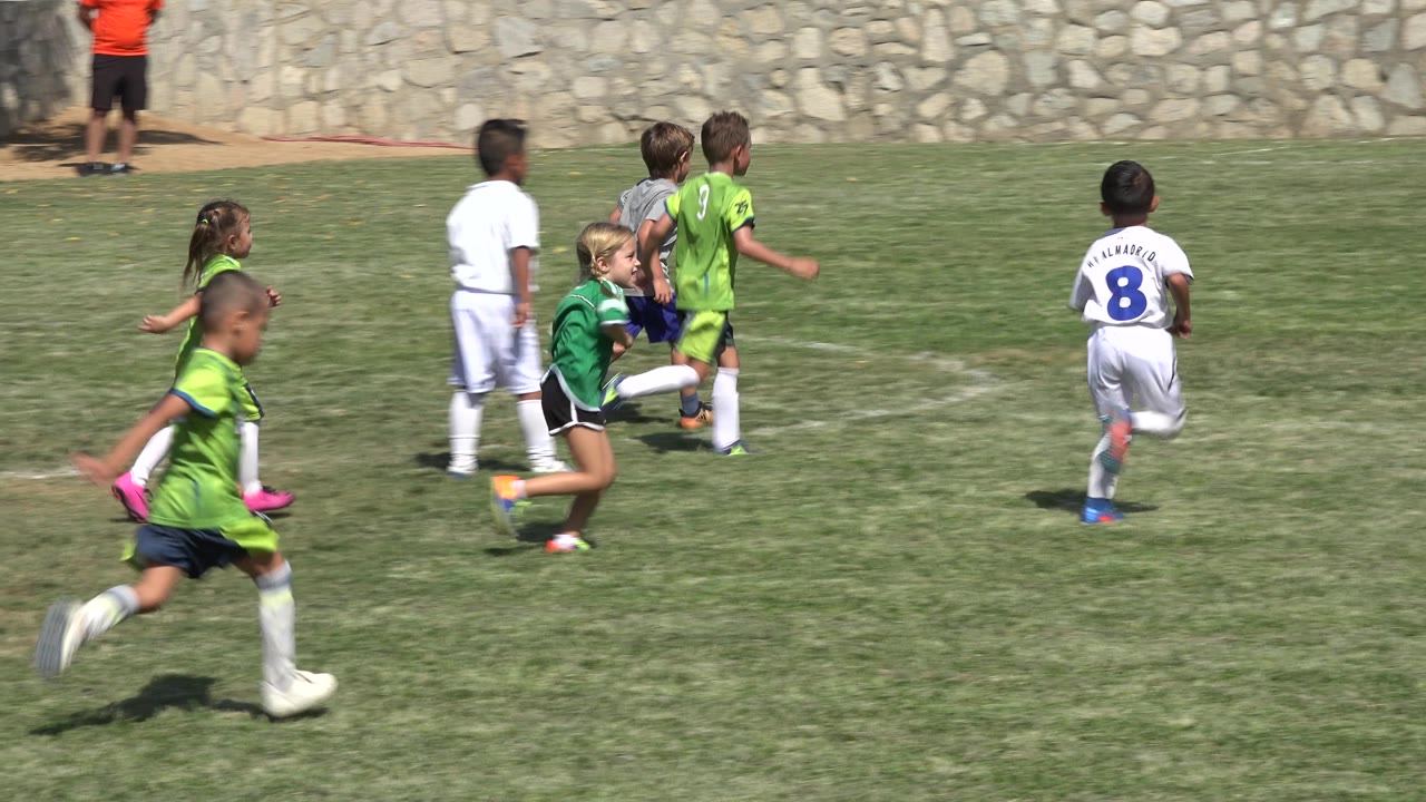 Several children continue running across the grass soccer field in San Jose del Cabo, Mexico, with green jerseys nearest the camera and white uniforms farther back.
