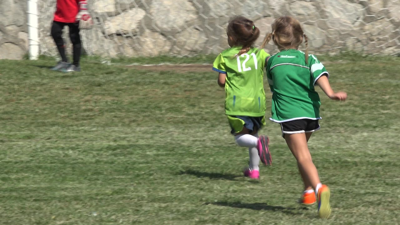 Two young children in green soccer jerseys run across a grass field near a stone wall.