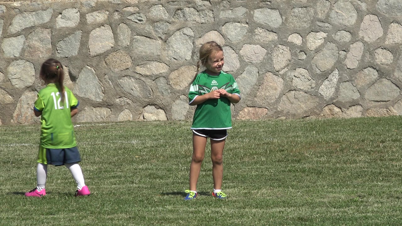 Two children in green soccer jerseys on a grass field by a stone wall.